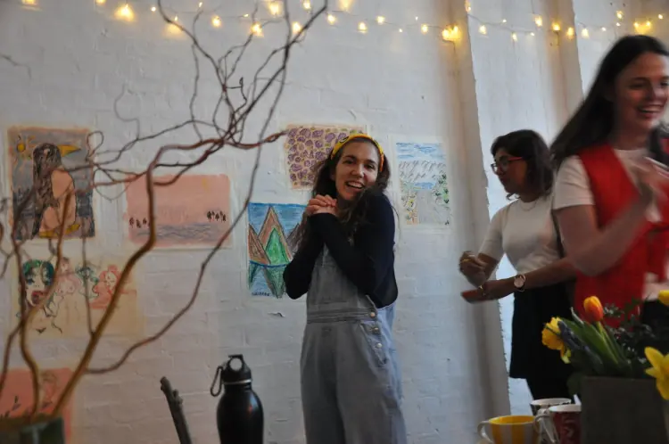 A guest laughing in front of a brick wall covered in colourful drawings, under string lights, with other guests chatting behind them
