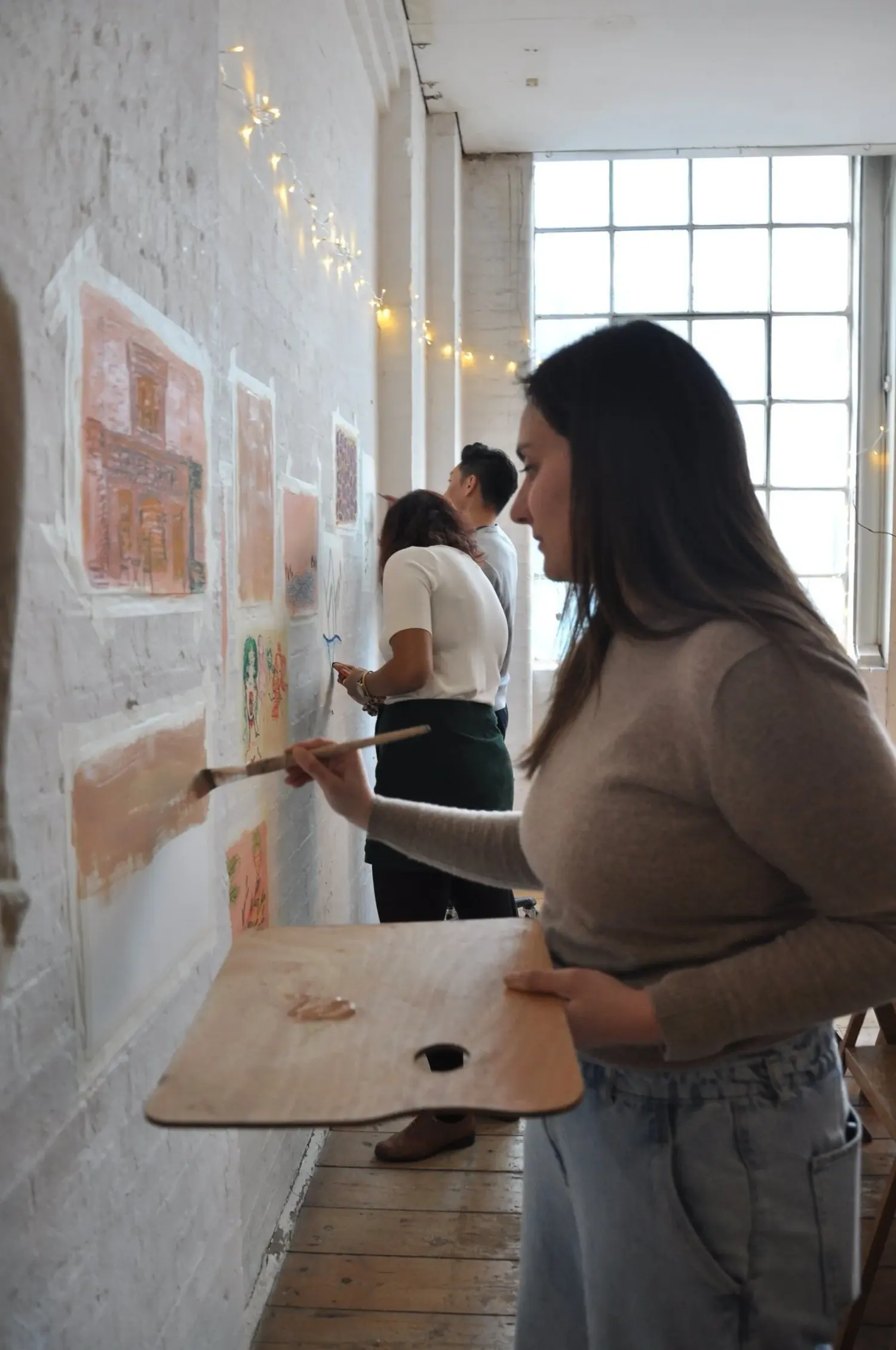 A guest painting on the community wall with a large brush and a wooden palette, while other guests paint further along the white brick wall