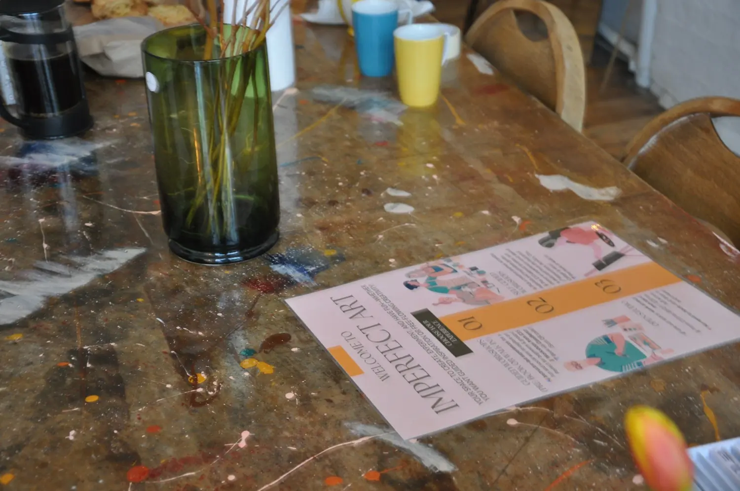 A paint-splattered wooden table with a French press, coloured mugs, a vase of dried grasses and a Welcome to Imperfect Art leaflet
